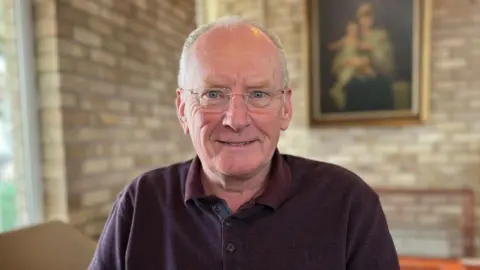 Brendan Flaxman is sat in a chair in a brick room with a window to the left and painting on the wall to the right. He is wearing a maroon collared jumper and glasses and is smiling into the camera.