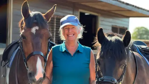 Karen Thompson standing on a yard between two bay horses with saddles and bridles on. She has blonde hair tied back and is wearing a purple cap and a blue, sleeveless polo top. There are wooden stables behind her. She is smiling at the camera. 