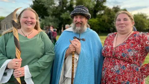 Three druids smile at the camera - two women, one man. Two hold wooden staffs and one in the middle wears a long blue hooded robe.