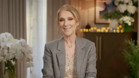 Céline Dion sitting on a chair with flowers behind her, smiling and wearing a grey blazer with a cream top