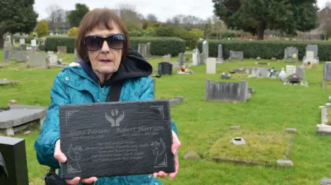 Edward Barnes Gina Jacobs, wearing sunglasses and a green coat, holds up a memorial plaque to her dead son at a cemetery