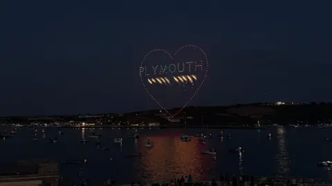 Visit Plymouth Colourful star-shaped drone lights displayed in the sky above a harbour with boats.