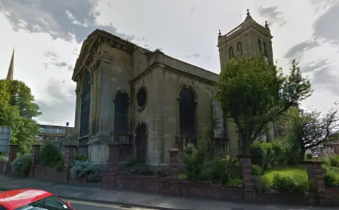 A stone church with a tower and large windows, with trees and a grass area outside. To the left, a spire from a different church can be seen. Part of a red car can be seen in front of the church. 