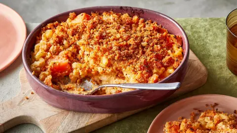 A pink bowl with mac and cheese and a crumbed topping. A spoon sits in the side.