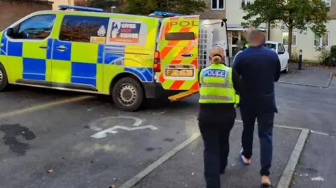 Hertfordshire Constabulary A police offer leading a man to a police vehicle. The man is dressed in dark clothing and is wearing sliders. The officer is female, with her hair in a low bun and is wearing a police uniform. There are parking spaces, one with a disabled sign, and flats in the distance with a white car in front and trees. 