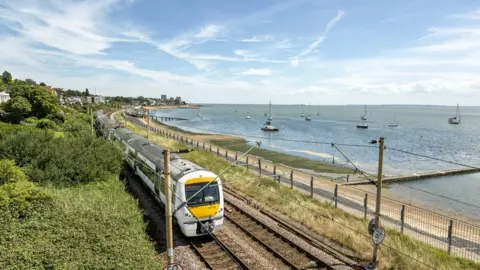 Andy Rose A white, yellow and grey c2c train travels on the railway beside a stretch of sandy beach and sea on one side, and a grassy bank on the other. It is a sunny day. There are blue skies.