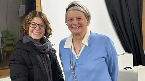 Sarah Jones (left) with brown bobbed hair wearing black glasses and a black coat stands with Angela Ralph (right) who has grey hair short hair with a black headband wearing a blue cardigan and glasses round her neck. They are both smiling and standing in the community room.
