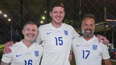 Karl Duguid Jamie Cureton, Greg Halford and Karl Duguid, all wearing a white England top, are standing with their arms around each other by a floodlit football pitch at night.