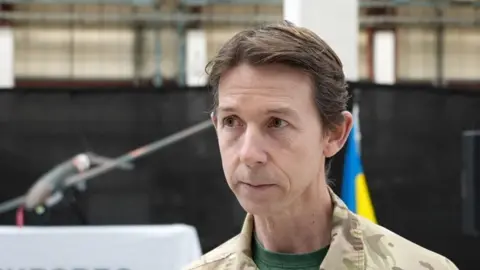 Shaun Whitmore/BBC A head and shoulders image of Colonel Andy Boardman. He is wearing a camouflage coloured shirt over a green T-Shirt. He is looking just past the camera. 