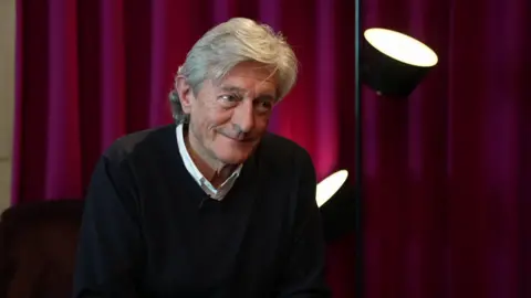 Nigel Havers sits in front of a burgundy curtain with lights in front of it.