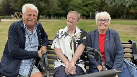 A man with short grey hair and a moustache in a blue shirt and jacket leaning next to a park bench. On the bench is a man smiling in a black and white shirt with a woman sat next to him with glasses and short blonde hair.