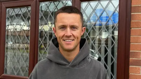 Stuart Dunn Stuart Dunn has short brown hair and is wearing a grey hoodie. He is stood outside a house and smiling, looking at the camera.