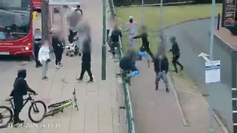 A group of masked young people at a bus stop. One appears to be carrying in a weapon. 