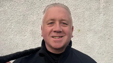 A close-up image of Bernie Leddy. He is an older man with long, grey hair which is tied back. He is standing in front of a white wall and is smiling at the camera. On his shoulder, he is holding a shillelagh - a dark stick used in Irish stick fighting.