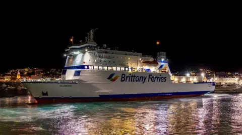 Brittany Ferries A Brittany Ferries ferry docked in St Peter Port in Jersey at night. The boat is lit up by lights with the lights reflecting on the water. The boat is white with red and blue on it along with the company name and logo. 