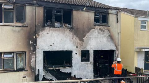 A person in an orange hi-vis jacket takes a photo of the badly damaged terraced house, with charred and broken windows and front door and smoke and fire damage visible