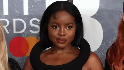 Sugababes at the Brit awards red carpet. Keisha is in the centre, wearing a black dress with a statement collar like a hoop round her neck