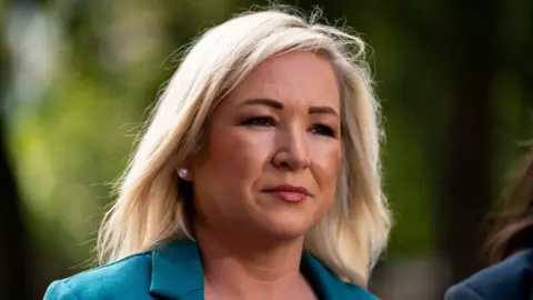 PA Media Michelle O'Neill - a woman with blonde, shoulder-length hair looks beyond the camera. She is wearing a green suit jacket. The background is blurred.