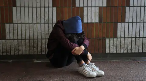 PA A masked teenage girl sits in front of a tiled wall in a pedestrian underpass with her head resting on her knees