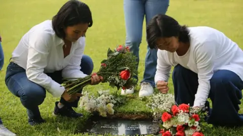 GERMAN FALCON/EPA/Shutterstock Presidential candidate Keiko Fujimori (left) visits the grave of her father, former Peruvian President Alberto Fujimori, at the Campo Fe Cemetery accompanied by her sister Sachi (R), in Lima, Peru, 12 April 2026. Both sisters are wearing jeans and white long-sleeved T-shirts. They are laying flowers on the grave.  
