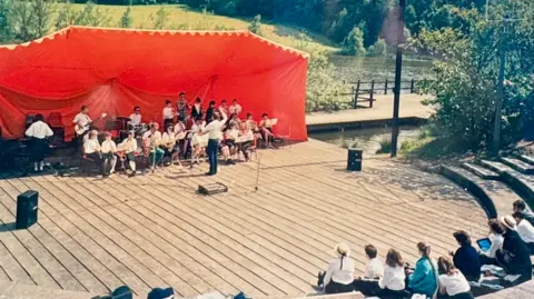 Matt Pearson A large wooden stage with performers and a large red covering behind the stage and a few people sitting on steps in front of it