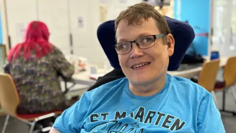 A man in his mid 40s in a wheelchair. he is wearing a sky blue top and wears black rimmed glasses. 
