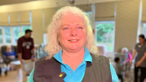 A woman with blonde hair is smiling for the camera. She is wearing a blue polo shirt and a dark grey gilet. She is standing in room with elderly people in the background.