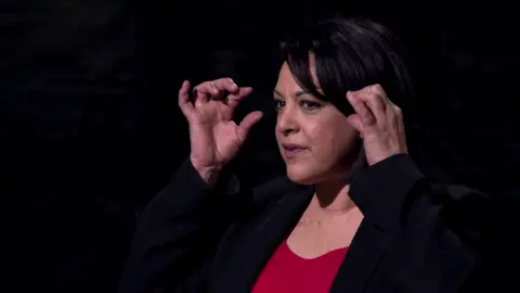Noora is standing before the Dragons, who cannot be seen, wearing a dark jacket and red top. She is gesturing with her hands around her eye area.