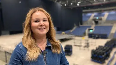 Rhiannon Newman from Culture Liverpool stands at a venue wearing a denim dress.