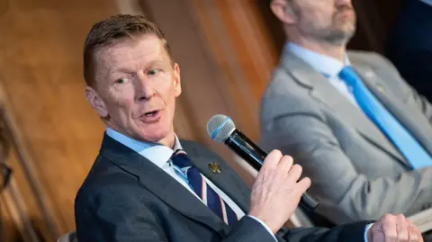 Tim Peake, who has ginger hair and is wearing a suit and blue and white tie, is pictured holding a microphone