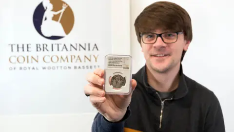 Britannia Coin Company Christopher Collects, wearing black top and glasses, holding the coin in front of sign saying 'The Britannia Coin Company'.