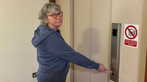 An elderly women standing in front of a closed lift door, pressing the call button. There is a sign on the wall next to the elevator with a red circle and diagonal line which says not to use the lift in the event of a fire. She is wearing a blue jumper and wearing glasses. 