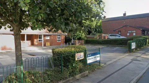 A red-brick building behind a large concrete car park, surrounded by green fencing. 