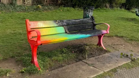 NORTH HERTS PRIDE A park bench painted in various colours which has been set on fire as half of it is blackened.