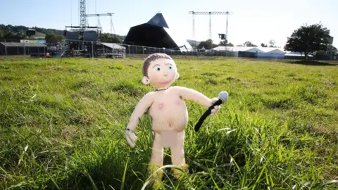 Jason Bryant A knitted doll resembling Chris Martin standing on the grass in front of Pyramid Stage at Glastonbury. The doll is naked and holding a microphone