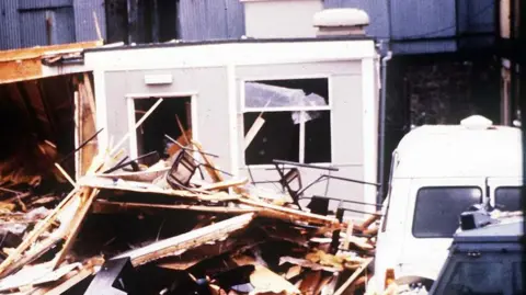 Pacemaker The aftermath of a bomb.  Wood and debris from the base piled up on the ground and a range rover dark blue car with an officer dressed in black standing in front of the scene. 