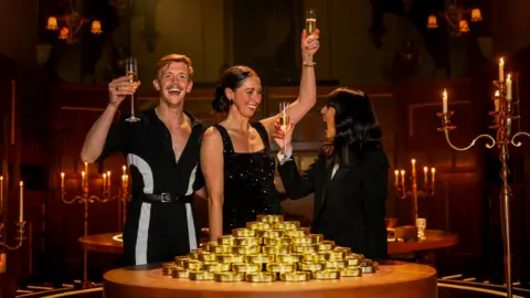 The jubilant winners of the third series of The Traitors - Stephen Libby and Rachel Duffy - hold up glasses of champagne behind a pile of gold coins. Host Claudia Winkleman toasts them with a glass of champagne. 