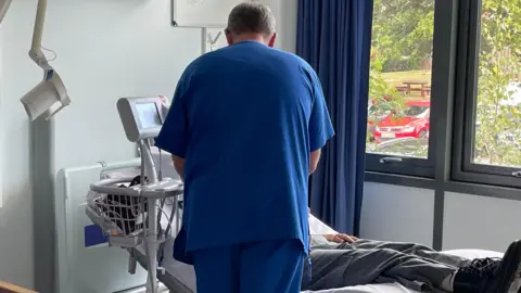 Man facing away from camera wearing dark blue scrubs looking down at a person on a hospital bed fully clothed on top of the bed wearing grey cargos and black boots, monitor by the side of the bed with a window looking out onto a car park