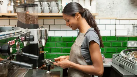 Female barista with long dark hair in a ponytail standing in front of a coffee machine in a kitchen with green and white tiles looking at her phone