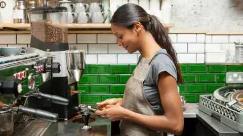 Getty Images Female barista with long dark hair in a ponytail standing in front of a coffee machine in a kitchen with green and white tiles looking at her phone