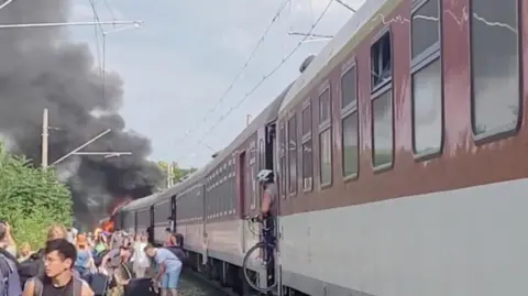 Reuters Passengers walk away from a train that is on fire at one end, with one man leaving the train with his bicycle.