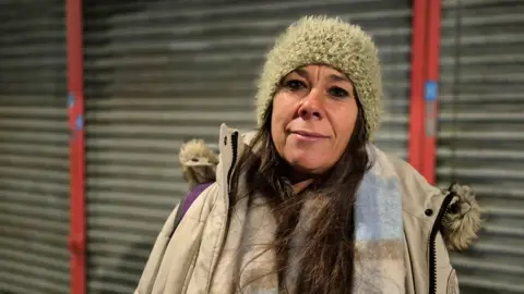 A woman with brown hair and wearing a gold-coloured furry hat and beige winter coat and scarf stands on a street on a dark night with grey shop shutters behind her. She is looking at the camera with half a smile.
