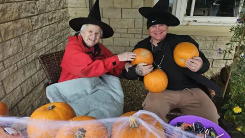 Catherine House Care Home A woman wearing a black witches hat is sitting next to a man holding three pumpkins. They are both sitting in front of a table with pumpkins and sweets on it.