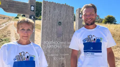 Alex and Miles standing by a sign which says Eastbourne to Winchester 100 miles. Both are wearing t-shirts for their charity.