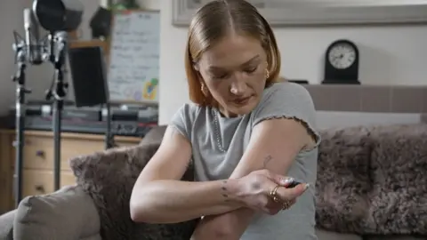 Anna is pictured sitting on a grey sofa in her living room. She is injecting herself with an insulin pen. Anna is wearing a grey T-shirt and she has strawberry blonde hair. Over her left shoulder is a microphone on a stand, some DJ decks and a small whiteboard. 