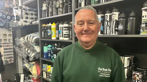 Chris Tate, standing in The Back Page shop in front of shelves of Newcastle United memorabilia. He has grey hair and is smiling at the camera. He is wearing a green jumper with the shop's name written on it.