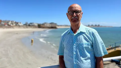 Joao stands on a beachside boardwalk, wearing a light blue patterned short-sleeve shirt. Behind them are a sandy beach, calm ocean waves, and houses along the coastline under a clear blue sky.