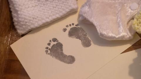 A piece of white paper with dark baby footprints lies next to baby blankets on a dark wooden table top