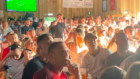 Angela Ferguson/BBC Fans wearing England tops are sitting at long tables and standing inside a large barn.