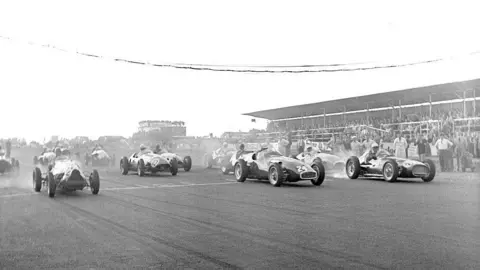 An old black and white picture of 1950s race cars setting off on a race at Castle Combe circuit. There is a spectator stand in the background, partly sheltered at the top. More buildings can be seen in the distance.